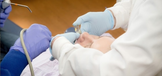 Dentist in white coat holding blue dental tool examining patient's mouth in clinical setting.