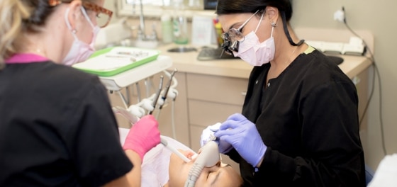 Dental professional in black mask and purple gloves examines patient's teeth in clinical treatment room.