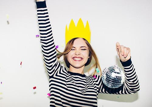 Child wearing striped shirt and yellow crown celebrating with confetti against white background.