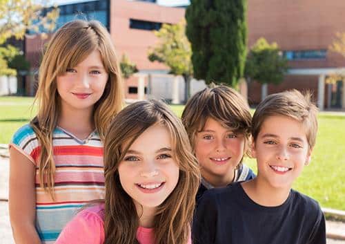 Four smiling children standing together outdoors on a sunny school playground lawn.
