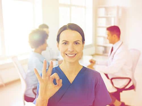 Female dental professional in scrubs giving okay sign in modern dental office with colleagues in background.