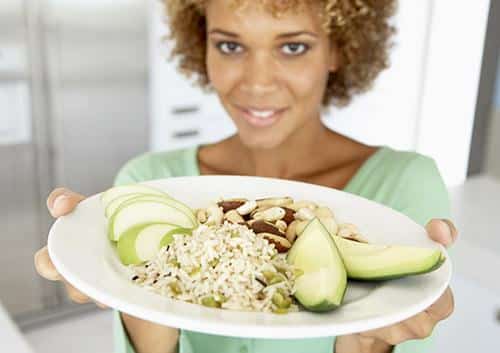 Woman in green shirt holding plate of healthy food with avocado and grains, smiling at camera.