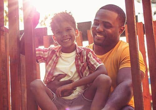 Man and young child smiling together outdoors in warm sunlight, wearing casual clothing.