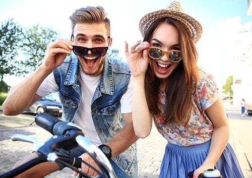 Two young people wearing sunglasses smile while holding a bicycle outdoors on a sunny day.