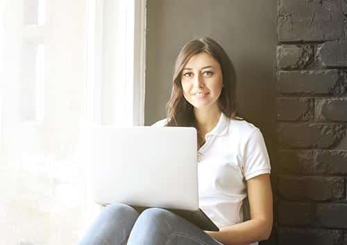 Woman in white shirt using laptop while sitting against brick wall near window.