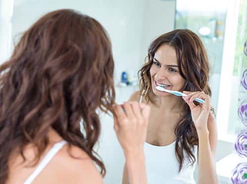 Woman brushing teeth at bathroom mirror with toothbrush and smile.