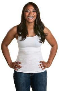 Smiling woman in white tank top and jeans poses with hands on hips against white background.