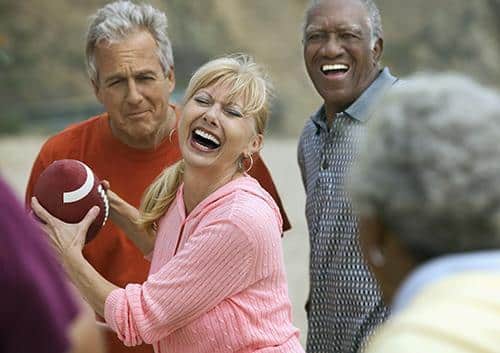 Three older adults laughing and playing football outdoors on a sunny day.