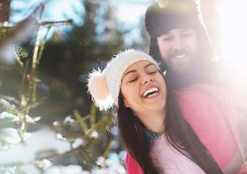 Woman wearing white winter hat smiling outdoors near Christmas tree with snow.