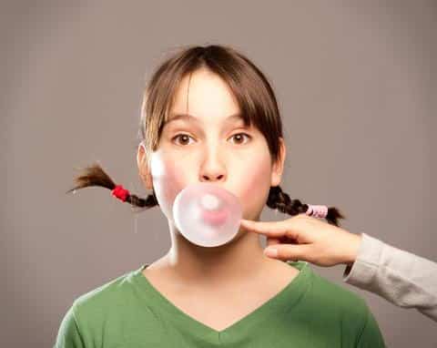 Young person blowing pink bubble gum against neutral gray background.