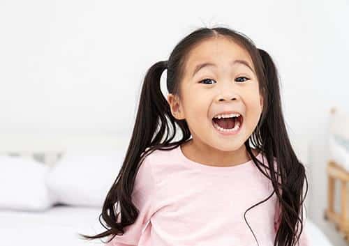 Young child with long dark hair smiling brightly in pink shirt against white background.
