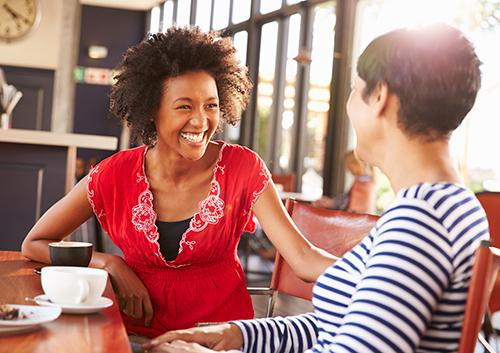 Two women smiling while having coffee together at a wooden table indoors.
