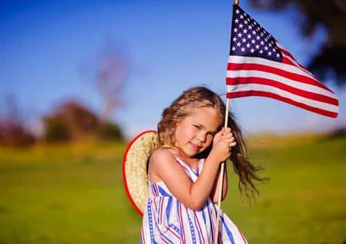 Young girl holding American flag in sunny outdoor field, smiling at camera.