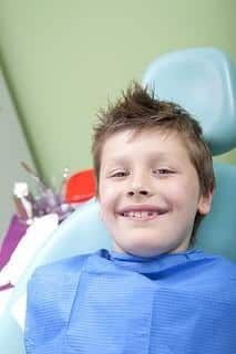 Young boy smiling in dental chair wearing blue bib at pediatric dental office.