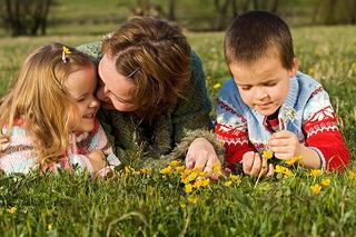 Three young children lying on grass, picking yellow flowers outdoors in a field.
