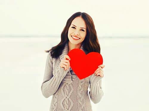 Woman holding red heart shape, smiling at camera, neutral background, dental/healthcare context.