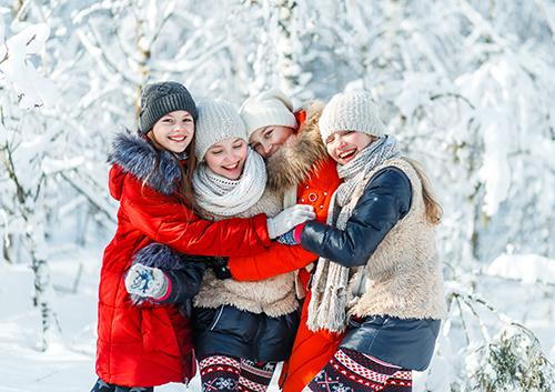 Family of four in winter clothing hugging outdoors in snowy forest setting.