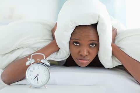 Woman in bed covering head with pillow next to alarm clock, appearing tired or frustrated.