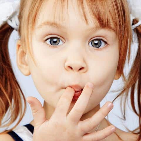 Young child with reddish-brown hair touching mouth with finger against white background.