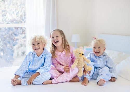 Three young children sitting together on a bed with a stuffed bear toy.