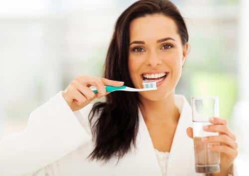 Woman brushing teeth with glass of water in bright indoor setting, smiling.