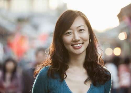 Woman smiling at camera in blue shirt, blurred urban street setting in background.