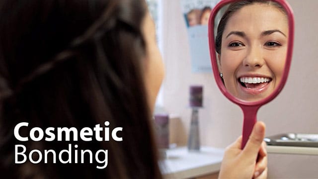 Woman examining smile in handheld mirror in dental office bathroom setting.