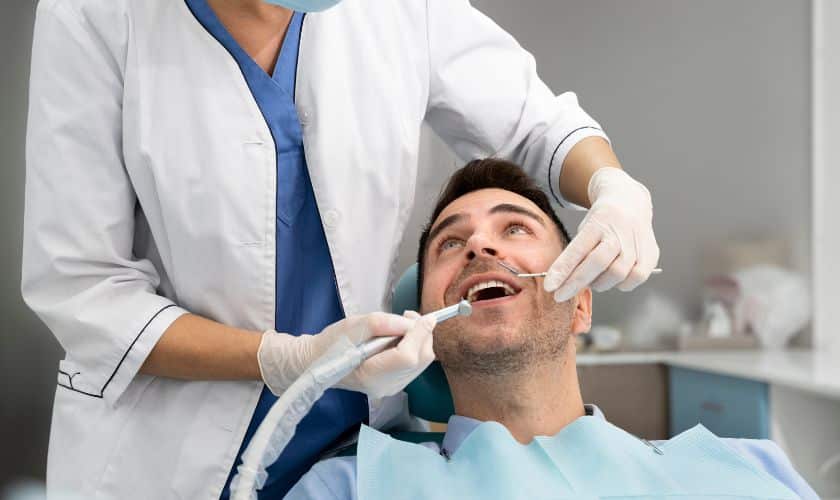 Dentist in white coat examining male patient's teeth in clinical dental office setting.