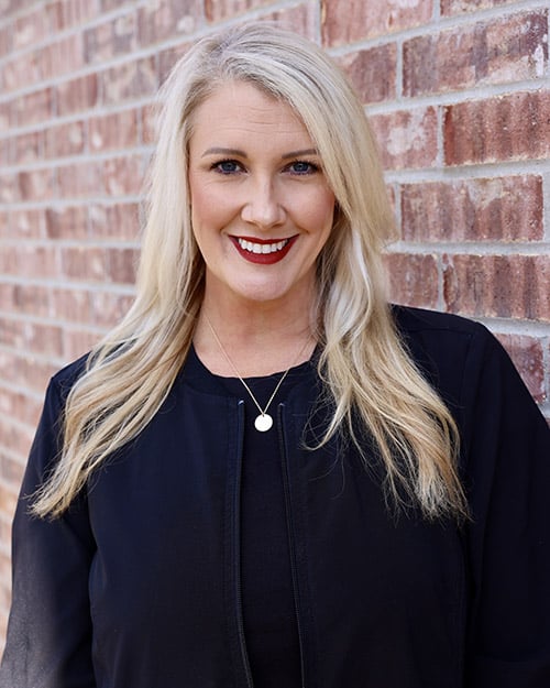 Professional headshot of smiling woman with long blonde hair against brick wall background.
