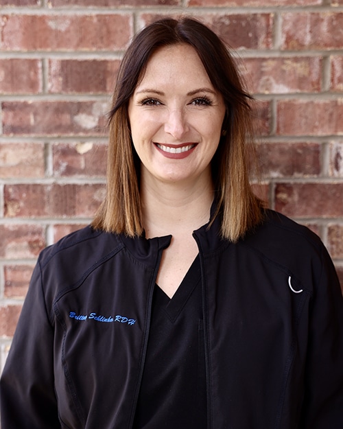 Professional woman smiling at camera against brick wall, wearing black dental scrubs.