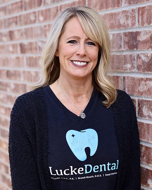 Woman smiling at camera wearing LuckeDental branded black shirt against brick wall background.