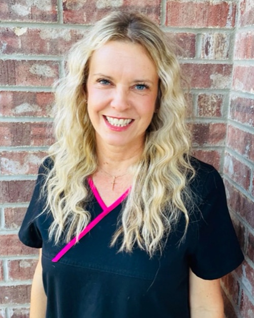 Woman smiling at camera wearing black shirt with pink accent against brick wall background.