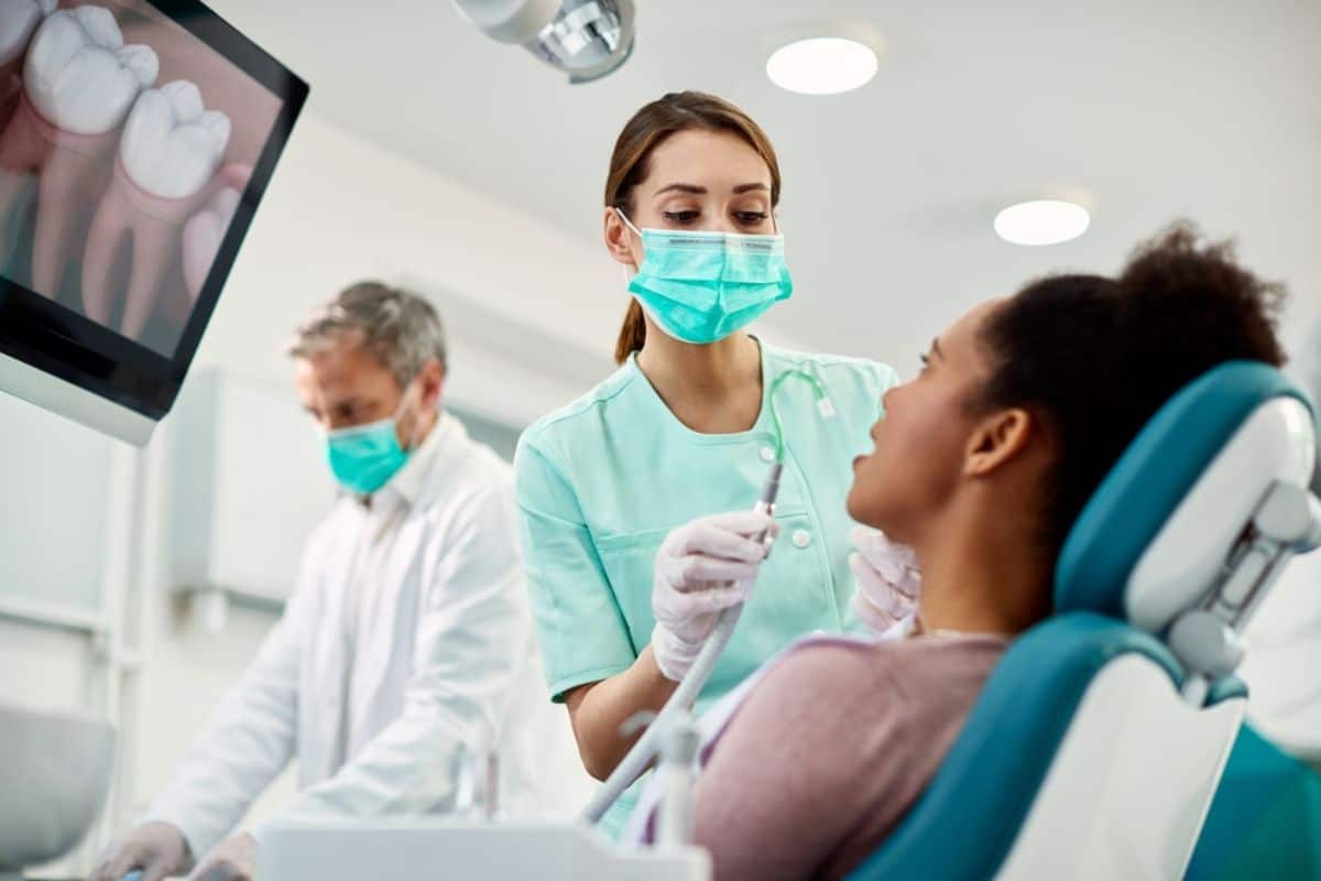 Dental hygienist in mask assists patient in treatment chair while dentist observes in modern clinic.