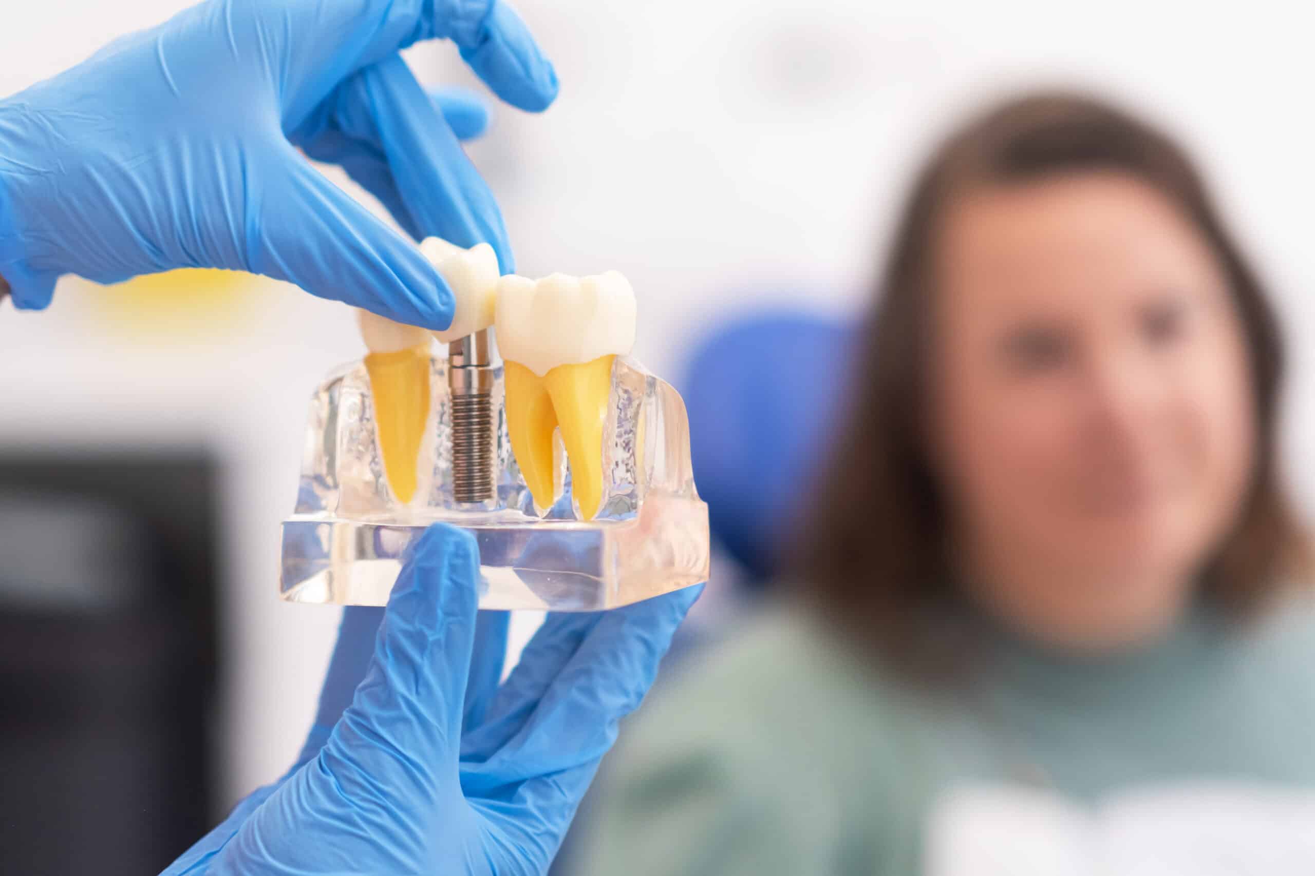 Dentist in blue gloves holding extracted tooth specimen with patient visible in blurred background.
