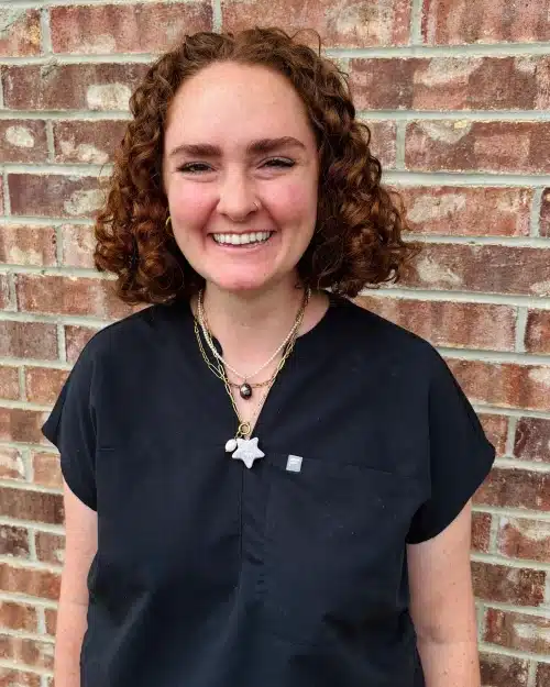 Smiling woman with curly brown hair wearing dark scrubs against brick wall background.