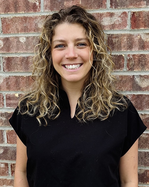 Woman smiling at camera against brick wall, wearing black shirt for dental practice headshot.