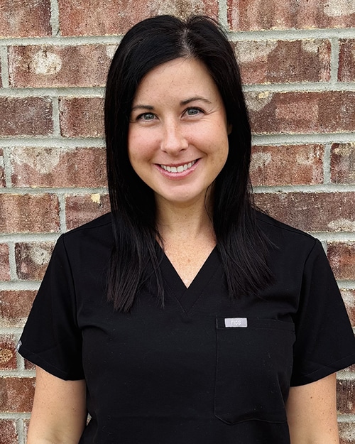 Woman in black scrubs smiling at camera against brick wall background.