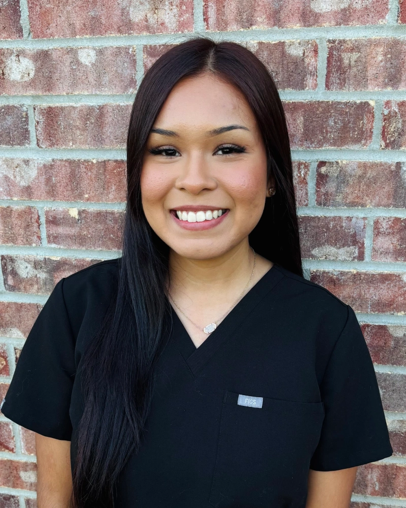 Young woman smiling at camera wearing black scrubs against brick wall background.
