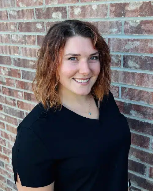 Young woman with reddish-brown wavy hair smiling at camera against brick wall background.