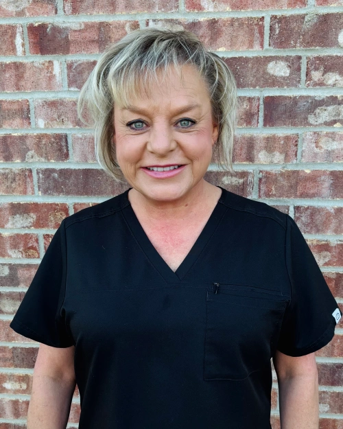 Professional woman in black scrubs smiling at camera against brick wall background.