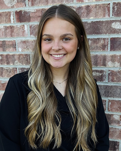 Young woman smiling at camera against brick wall background, professional headshot style photo.