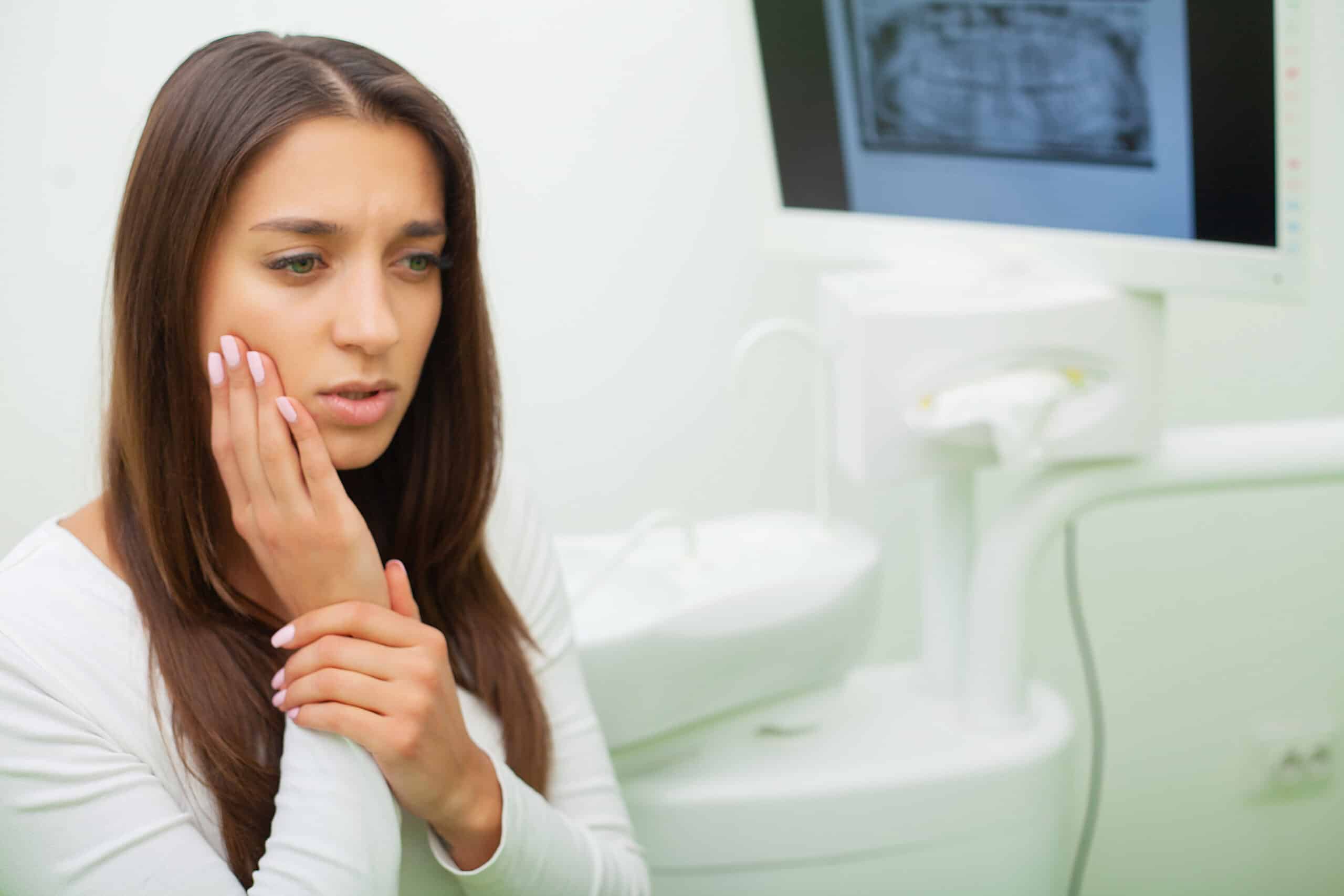 Woman touching her cheek in pain at dental office with equipment visible in background.